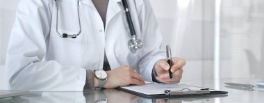 Klinik muayenesi sırasında panoya steteskopla yazı yazan beyaz önlüklü doktor hasta notlarını ve reçeteli verileri inceliyor. İlaç ve sağlık hizmetleri