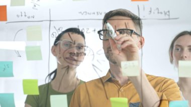 İş arkadaşları beyin fırtınası yapıyor, cam tahtaya strateji notları yazıyor, yaratıcı iş yeri toplantısında renkli yapışkan notlarla işbirliği yapıyorlar. Takım çalışması konsepti.