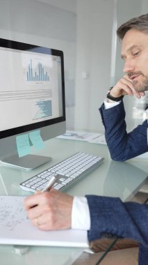 Takım elbiseli işadamı masa başına odaklanmış, klavyeye yazarken aynı zamanda kalemle not alıyor, modern ofis ortamında birden fazla iş yapıyor. İş adamları konsepti