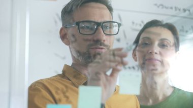 Professional team members brainstorming strategy, writing notes on transparent whiteboard with colorful sticky papers during collaborative meeting in office. Business people at work.