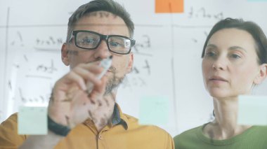 Two business colleagues are analyzing notes on a transparent whiteboard during a brainstorming session, working together to develop innovative solutions and strategies for their company