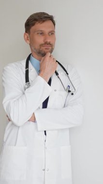 Kendine güvenen erkek doktor kolları çapraz duruyor, steteskop ve beyaz önlük giyiyor. Düşünceli bir şekilde, tıp alanında profesyonelliği ve uzmanlığı somutlaştırıyor. İlaç ve sağlık hizmetleri