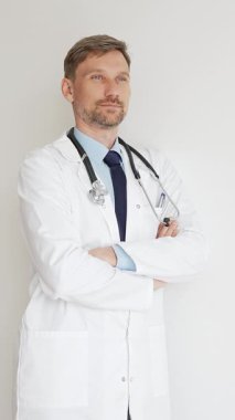 Kendine güvenen erkek doktor kolları çapraz duruyor, steteskop ve beyaz önlük giyiyor. Düşünceli bir şekilde, tıp alanında profesyonelliği ve uzmanlığı somutlaştırıyor. İlaç ve sağlık hizmetleri