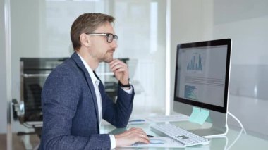 Profesyonel iş adamı gözlük takıyor ve spor ceket giyiyor, çağdaş bir ofis ortamında çalışıyor, bir masaüstü bilgisayarında görevler yapıyor. İş adamları konsepti