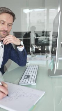 Takım elbiseli işadamı masa başına odaklanmış, klavyeye yazarken aynı zamanda kalemle not alıyor, modern ofis ortamında birden fazla iş yapıyor. İş adamları konsepti