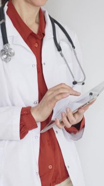 Tuğla kırmızısı bluz ve stetoskop üzerine beyaz önlük giyen kimliği belirsiz profesyonel doktor dijital tablet kullanıyor. Bu da tıpta yenilikçi sağlık uygulamaları olduğunu gösteriyor. Sağlık hizmeti kavramı