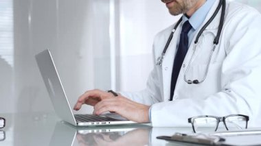Steteskoplu beyaz önlüklü bir doktor laptopta yazı yazıyor. Teletıp, sağlık ve dijital tıbbi kayıtları temsil ediyor. Tıp, sağlık ve bilim konsepti