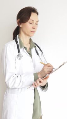 Steteskop takan doktor kadın ve yeşil bluzun üzerine beyaz önlük giymiş hasta bakımını ve tıbbi uzmanlığı temsil eden bir panoya yazı yazıyor. İlaç ve sağlık hizmetleri kavramı.