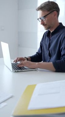 Focused businessman wearing glasses concentrating on his work, diligently typing on a laptop in a bright, modern office setting with a tablet and paperwork nearby. Business people concept.