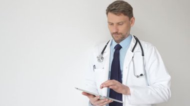 Beyaz önlüklü ve steteskoplu bir doktor dijital tablet kullanıyor. Hasta kayıtlarını güncellemeye odaklanmış, modern sağlık teknolojisi ve profesyonel uzmanlığın altını çizmiş. İlaç ve sağlık hizmetleri