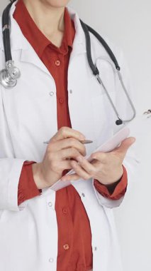 Tuğla kırmızı bluzun üzerine steteskop ve beyaz önlük giyen kimliği belirsiz kadın doktor tıbbi uzmanlığı ve hasta bakımını temsil eden bir panoya yazı yazıyor. İlaç ve sağlık hizmetleri kavramı.