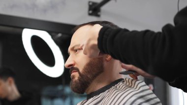Profesyonel berber elektrikli tıraş makinesi kullanıyor, modern bir berber dükkanında erkek bir müşterinin sakalını titizlikle kesiyor, şık ve parlak bir görünüm çiziyor.