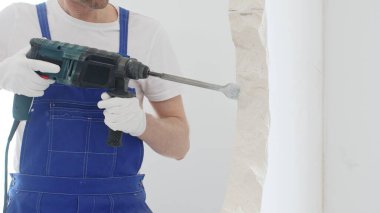 Mavi tulumlu ve koruyucu beyaz eldivenli tanımlanamayan erkek inşaat işçisi, beyaz duvarı döner çekiç matkapıyla yıkıyor, toz üretiyor ve yatay görüşü kapatıyor. Yenileme