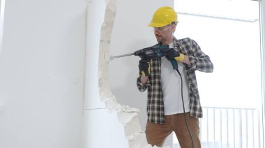 Bej kareli gömlek, sarı başlık ve koruyucu eldiven giyen erkek inşaat işçisi, dönen çekiç matkabıyla beyaz duvarı yıkıyor ve orta uzunlukta toz üretiyor. Yenileme projesi