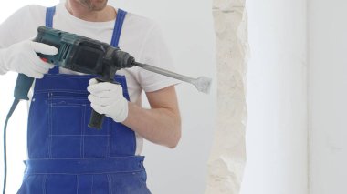 Mavi tulumlu ve koruyucu beyaz eldivenli tanımlanamayan erkek inşaat işçilerinin yakın çekiç matkabıyla beyaz duvarı yıkıyor, toz oluşturuyor, yatay görüş sağlıyor. Yenileme