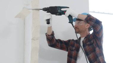 Kırmızı kareli gömlek, mavi başlık ve koruyucu eldiven giyen inşaat işçisi, döner çekiç matkabıyla beyaz duvarı yıkıyor, toz ve portre görüntüsü yaratıyor. Yenileme konsepti.
