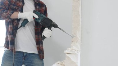 Kırmızı kareli kotlar ve koruyucu eldivenler giyen kimliği belirsiz erkek inşaat işçilerinin yakın çekiç matkabıyla beyaz duvarı yıkıp toz oluşturuyor. Yenileme konsepti.