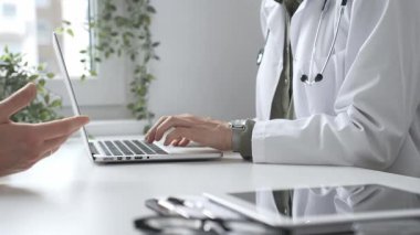 Klinik ortamdaki sağlık hizmetleri sırasında hasta bilgilerini, laptopunu ve steteskopunu kayıt altına alan beyaz önlüklü bir tıbbi uzman. İlaç ve sağlık hizmetleri kavramı