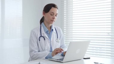 Laboratuvar önlüğü ve steteskop giyen kendine güvenen kadın doktor, doğal ışık dolu, parlak ve modern bir muayenehanede çalışırken laptopuna odaklanmış bir şekilde bir masada oturuyor.
