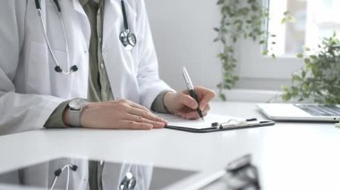 Laboratuvar önlüğü ve steteskop giymiş kadın doktor, tıp bürosunun masasındaki panoya profesyonelliği vurgulayan dizüstü bilgisayar ve gözlüklerle notlar yazıyor. İlaç ve sağlık hizmetleri kavramı