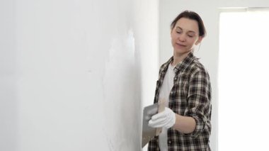 Kareli gömlek giyen kadın inşaat işçisi, beyaz duvar yüzeyini düzleştiriyor, profesyonel spatula tekniği ile macunu uyguluyor, iç dekorasyon konusunda yetenekli..