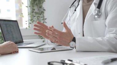Laboratuvar önlüğü ve steteskop giymiş bir erkek doktor modern tıp ofisinde tablet kullanan hastaya teşhis koymayı açıklarken el kol hareketi yapıyor. İlaç ve sağlık hizmetleri kavramı.