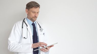 Beyaz önlüklü ve steteskoplu bir doktor dijital tablet kullanıyor. Hasta kayıtlarını güncellemeye odaklanmış, modern sağlık teknolojisi ve profesyonel uzmanlığın altını çizmiş. İlaç ve sağlık hizmetleri