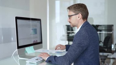 Profesyonel iş adamı gözlük takıyor ve spor ceket giyiyor, çağdaş bir ofis ortamında çalışıyor, bir masaüstü bilgisayarında görevler yapıyor. İş adamları konsepti