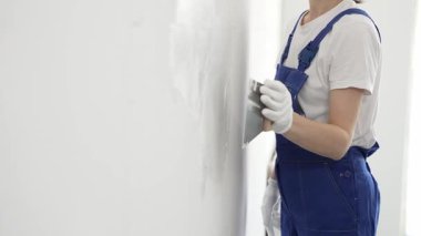 Meçhul inşaat işçisi beyaz eldiven ve mavi tulum giyen kadın, macun bıçağı kullanarak beyaz bir duvara alçı sürüyor. Ev yenileme projesi sırasında pürüzsüz bir yüzey oluşturuyor.