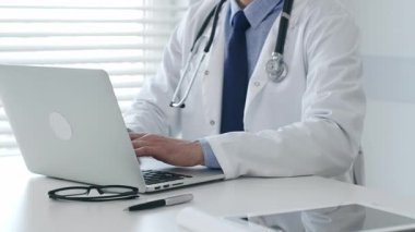 Laboratuvar önlüğü ve steteskop giyen tıp doktoru masasında oturuyor ve güneş gözlüğü ve kalemle yakın bir yerde, parlak bir tıp ofisinde dizüstü bilgisayarda yazıyor. İlaç ve sağlık hizmetleri kavramı