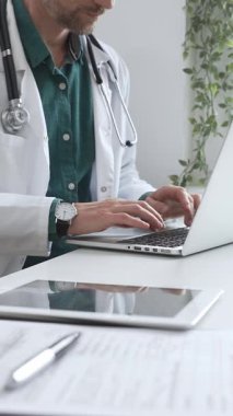 Doktor adam yeşil gömleğin üstüne beyaz önlük giyiyor ve steteskop da parlak bir tıbbi ofisteki dizüstü bilgisayarda yazarak sağlık ve teknolojinin kesişimini gösteriyor. İlaç ve sağlık hizmetleri.