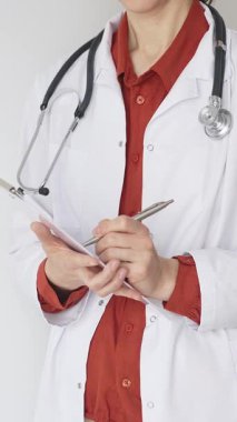 Tuğla kırmızı bluzun üzerine steteskop ve beyaz önlük giyen kimliği belirsiz kadın doktor tıbbi uzmanlığı ve hasta bakımını temsil eden bir panoya yazı yazıyor. İlaç ve sağlık hizmetleri kavramı.