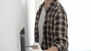 Koruyucu eldiven ve kareli tişört giyen kimliği belirsiz kadın inşaat işçisi, beyaz duvarda ıslak alçıyı düzleştiriyor ve iç dekorasyon sırasında profesyonel malayla malzeme yayıyor.