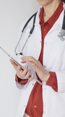 Tuğla kırmızısı bluz ve stetoskop üzerine beyaz önlük giyen kimliği belirsiz profesyonel doktor dijital tablet kullanıyor. Bu da tıpta yenilikçi sağlık uygulamaları olduğunu gösteriyor. Sağlık hizmeti kavramı