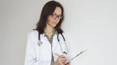 Yeşil bluzun üzerine steteskop, siyah gözlük ve beyaz önlük takan doktor kadın tıbbi uzmanlığı ve hasta bakımını temsil eden bir panoya yazı yazıyor. İlaç ve sağlık hizmetleri