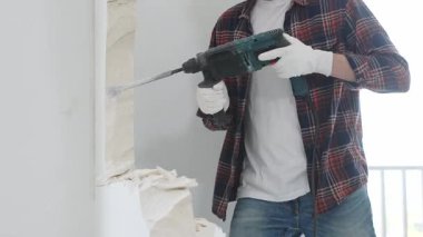 Kırmızı kareli kot pantolon ve koruyucu eldiven giyen kimliği belirsiz erkek inşaat işçisi, döner çekiç matkabıyla beyaz duvarı yıkıyor, toz üretiyor, yakın görüş sağlıyor. Yenileme konsepti.