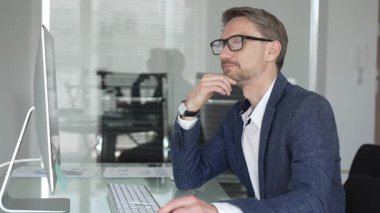 Profesyonel iş adamı gözlük takıyor ve spor ceket giyiyor, çağdaş bir ofis ortamında çalışıyor, bir masaüstü bilgisayarında görevler yapıyor. İş adamları konsepti