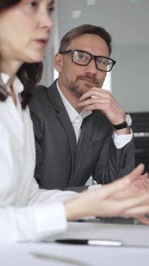 Takım elbiseli ve gözlüklü bir iş adamı önemli bir ofis toplantısında bayan bir meslektaşını dikkatle dinliyor, tartışma ve problem çözme üzerine yoğunlaşıyor, kurumsal strateji planlıyor.