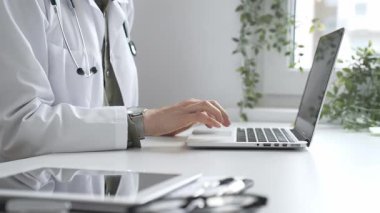 Stetoskop ve laboratuvar önlüğü giyen doktor özenle bir dizüstü bilgisayarda yazıyor ve modern bir tıp ofisinde verimli bir sağlık hizmeti yönetimi sergiliyor. İlaç ve sağlık hizmetleri kavramı