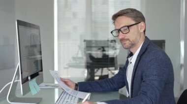 Modern ofis masasında gözlüklü orta yaşlı bir işadamı bilgisayar kullanırken belgeleri yazmaya ve analiz etmeye odaklanmış. İş adamları konsepti