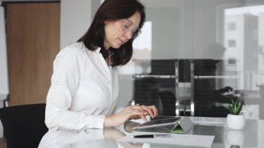 Modern cam bir masada dijital tabletle çalışan profesyonel bir ortamda çalışan kadın, üretkenliği, yeniliği ve teknolojinin kurumsal ofis ortamında entegrasyonunu vurguluyor.