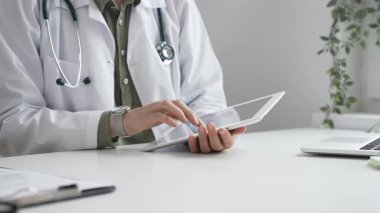 Beyaz laboratuvar önlüğü ve steteskop giyen profesyonel kadın doktor, klinik ortamdaki hasta kayıtlarını incelerken dijital tablet üzerinde çalışıyor. İlaç ve sağlık hizmetleri kavramı