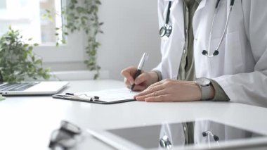Laboratuvar önlüğü ve steteskop giymiş kadın doktor, tıp bürosunun masasındaki panoya profesyonelliği vurgulayan dizüstü bilgisayar ve gözlüklerle notlar yazıyor. İlaç ve sağlık hizmetleri kavramı