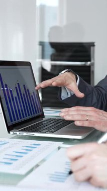 Bir dizüstü bilgisayarda iş birliği yapan ve mali tabloları inceleyen iş adamları, bir kişinin rapor üzerine notlar yazması, takım çalışması ve veri analizini göstermesi