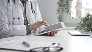 Beyaz laboratuvar önlüğü ve steteskop giyen profesyonel kadın doktor, klinik ortamdaki hasta kayıtlarını incelerken dijital tablet üzerinde çalışıyor. İlaç ve sağlık hizmetleri kavramı