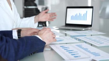 Ekonomi uzmanları finansal verileri analiz ediyor, pazar eğilimlerini tartışıyor ve bir dizüstü bilgisayar ekranındaki çizelge ve grafiklerle raporları gözden geçiriyor ve şirket planlama toplantısında basılı belgeler hazırlıyorlar.