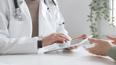 Laboratuvar önlüğü ve steteskop giymiş erkek doktor modern tıp ofisinde tablet kullanırken hastayı dinliyor. İlaç ve sağlık hizmetleri kavramı.