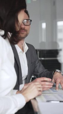 Kadın olarak masa başında işbirliği yapan iş adamları önemli bir sözleşme imzalarken, gözlüklü bir adam tablet üzerinde ayrıntılara dikkat çekiyor, takım çalışmasını ve karar verme mekanizmasını vurguluyor.