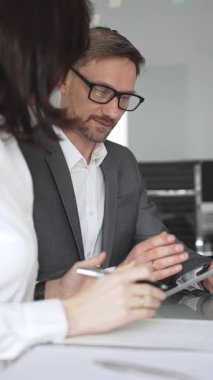 Kadın olarak masa başında işbirliği yapan iş adamları önemli bir sözleşme imzalarken, gözlüklü bir adam tablet üzerinde ayrıntılara dikkat çekiyor, takım çalışmasını ve karar verme mekanizmasını vurguluyor.