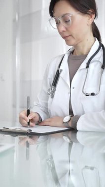 Beyaz önlüklü ve gözlüklü bir kadın doktor. Masada oturuyor, panoya yazıyor, hasta bilgilerini ve kayıtlarını yönetiyor. İlaç ve sağlık hizmetleri